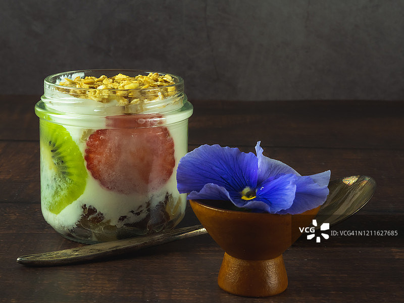 一杯含有猕猴桃、草莓和格兰诺拉麦片早餐的天然酸奶，一朵可食用的花，一把旧羊驼或银勺子放在木桌上图片素材
