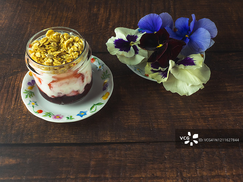 一杯加草莓、红莓果酱的原味酸奶和早餐格兰诺拉麦片，木桌上放一朵可食用的花图片素材