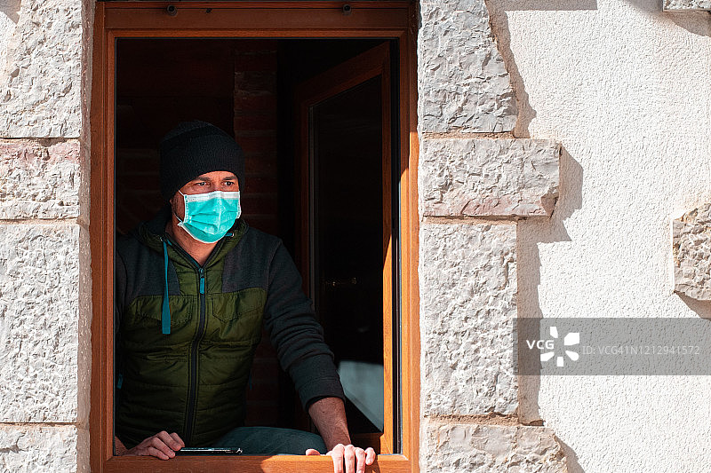 冠状病毒感染期间戴口罩的男子透过窗户向外看图片素材