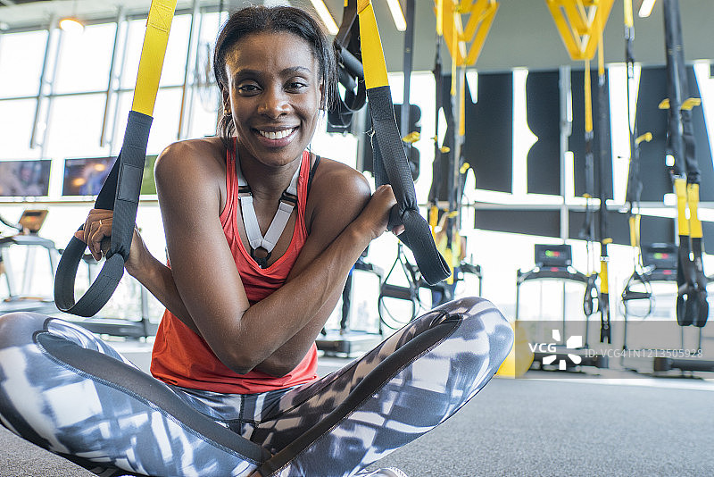 一个非洲裔哥伦比亚女人坐在健身房的地板上，手里拿着一个trx图片素材