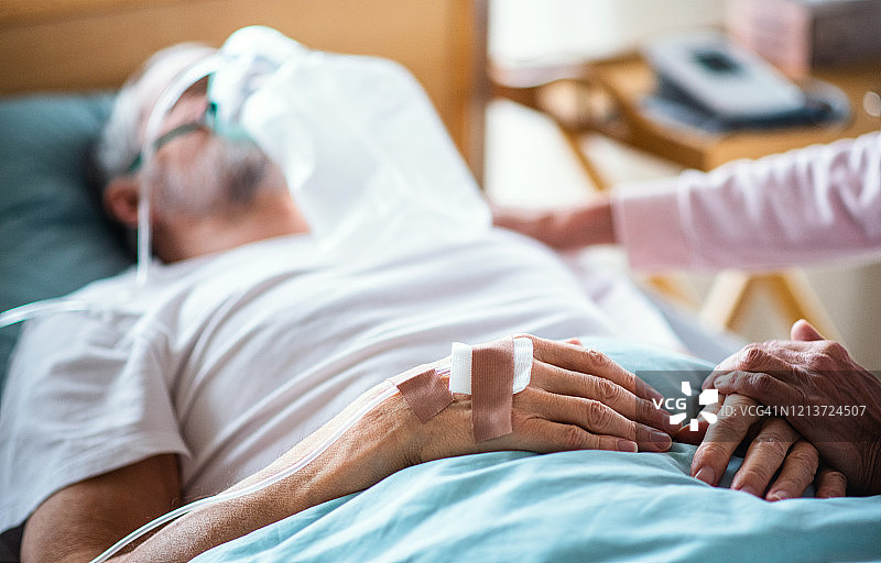 一位年老的病人躺在病床上，他的妻子牵着他的手。图片素材