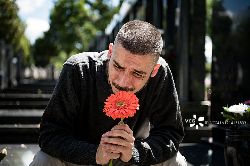 悲伤和沮丧的鳏夫在黑衣跪在墓碑前，拿着一朵花和哀悼家人的损失。死亡、哀悼、葬礼和灵性的概念。——Covid-19图片素材