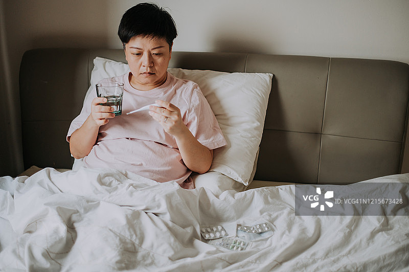 一位亚裔华裔女性在家生病，在床上用体温计测量体温，用水吃药图片素材