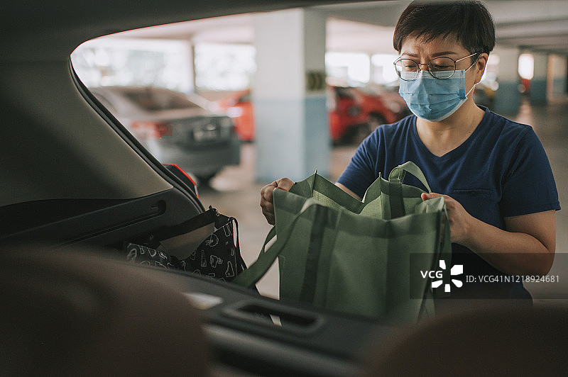 一位亚洲华人中年妇女从她的车里取出可重复使用的袋子，这些袋子是她刚从超市买的所有杂货，还带着口罩图片素材