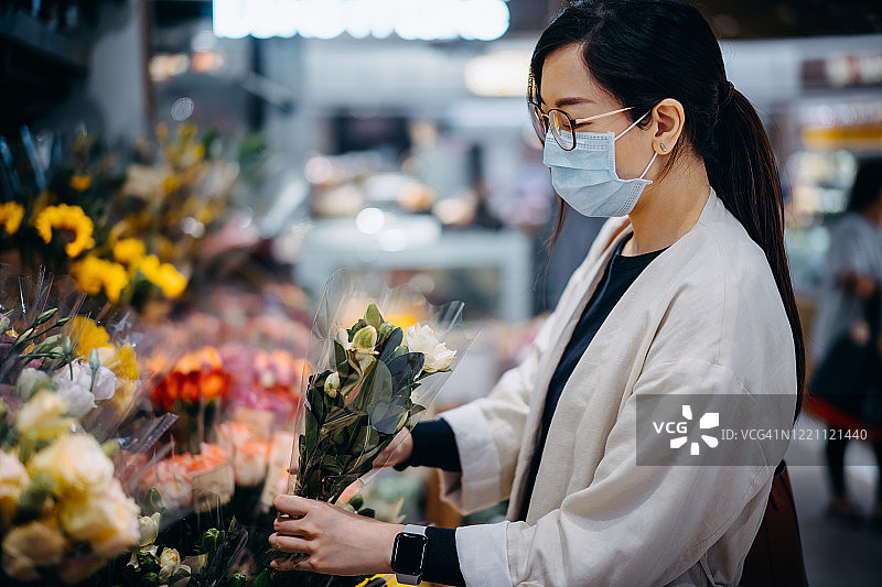 新冠肺炎疫情爆发期间，年轻的亚洲女性戴着防护口罩在花店购买鲜花图片素材