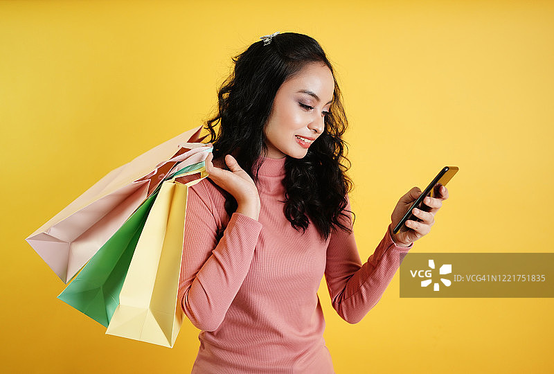 自信快乐的女人在网上购物，并通过手机应用订购产品。顾客女士手持购物袋孤立在黄色背景。移动应用技术概念。图片素材