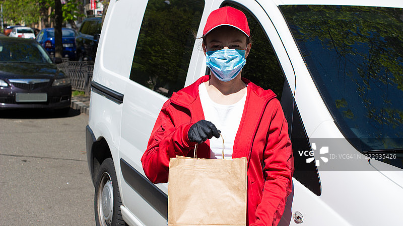年轻的女邮差在货物面前投递包裹。送餐女员工穿着红色制服，戴着防护口罩和医用手套，手持装有食品的牛皮纸包装袋。图片素材