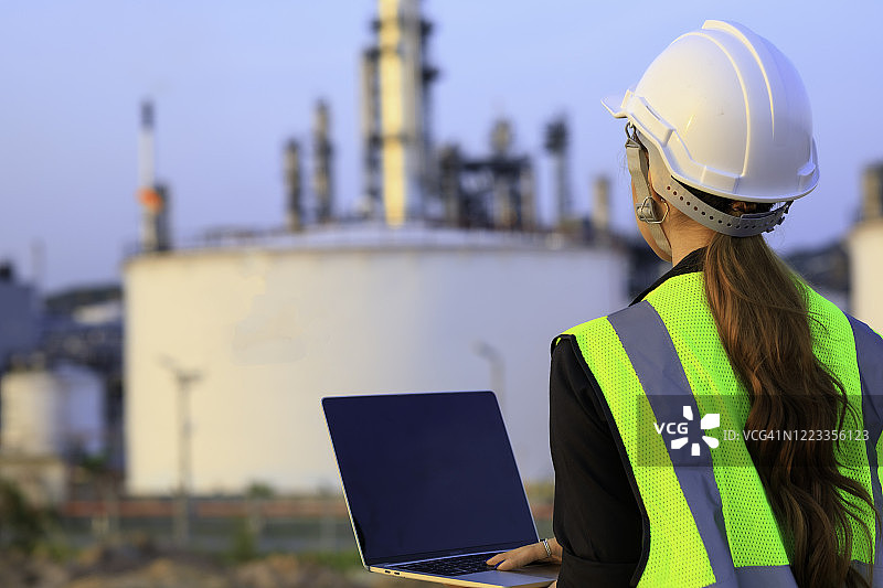 女工程师领班戴安全帽使用笔记本电脑在炼油厂工厂工作背景。图片素材