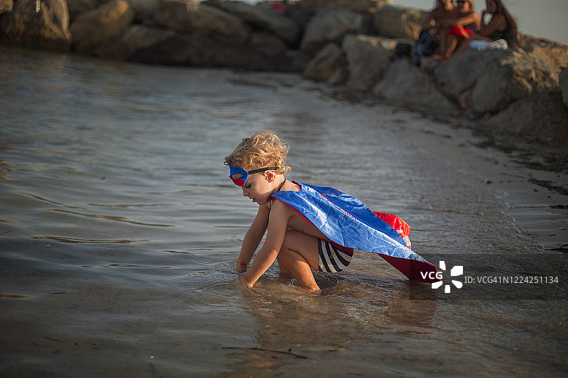 穿着泳衣和超级英雄服装的男孩在海滩上把手放在水里图片素材