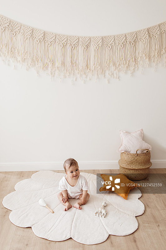 可爱的微笑蹒跚学步坐在贝壳地毯上。
可爱的婴儿与生态玩具，星星枕头和棉流苏坐在地板上美丽温柔的游戏室。图片素材
