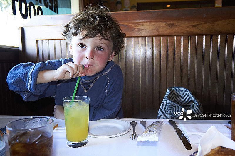 小男孩在一家餐馆的木棚里用绿吸管喝着一个大玻璃杯里的橙汁。图片素材