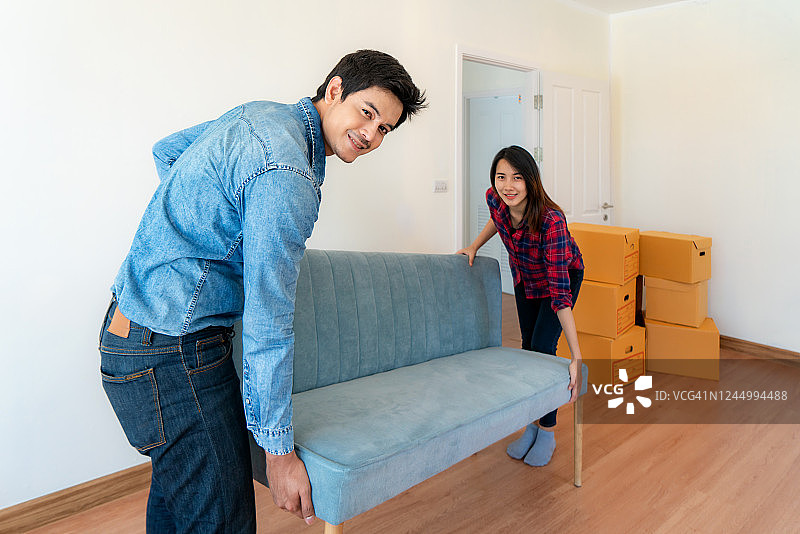 快乐的年轻亚洲夫妇举起沙发或沙发到卧室与纸板盒在他们的新家一起。搬家的日子。图片素材