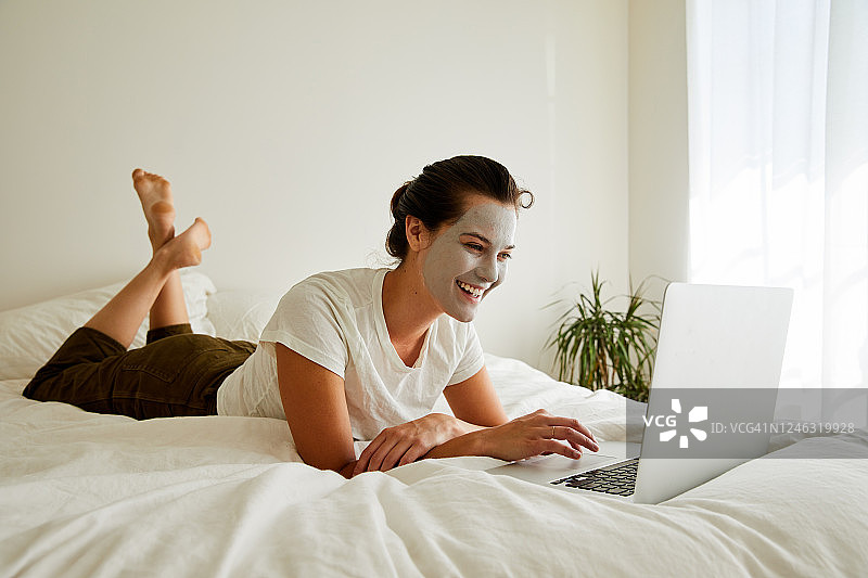 带着面具的笑女人躺在床上用笔记本电脑图片素材