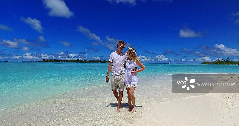 一对穿着白色婚纱的幸福夫妇在马尔代夫的热带海滩上散步。海浪在沙滩上翻滚图片素材