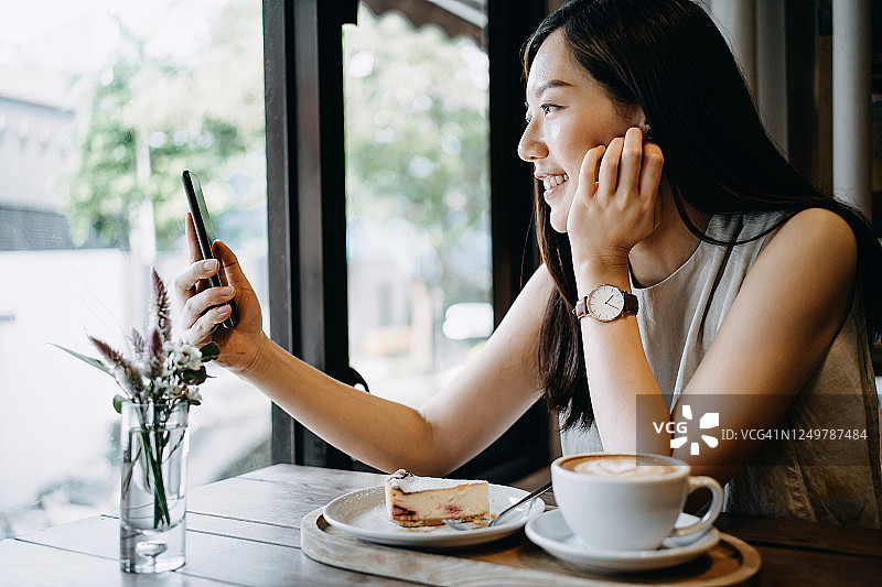 美丽微笑的年轻亚洲女子坐在咖啡馆享受一杯咖啡与蛋糕，并使用她的智能手机图片素材