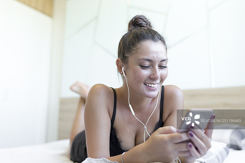 微笑的年轻女子享受音乐，同时使用智能手机在床上在家图片素材