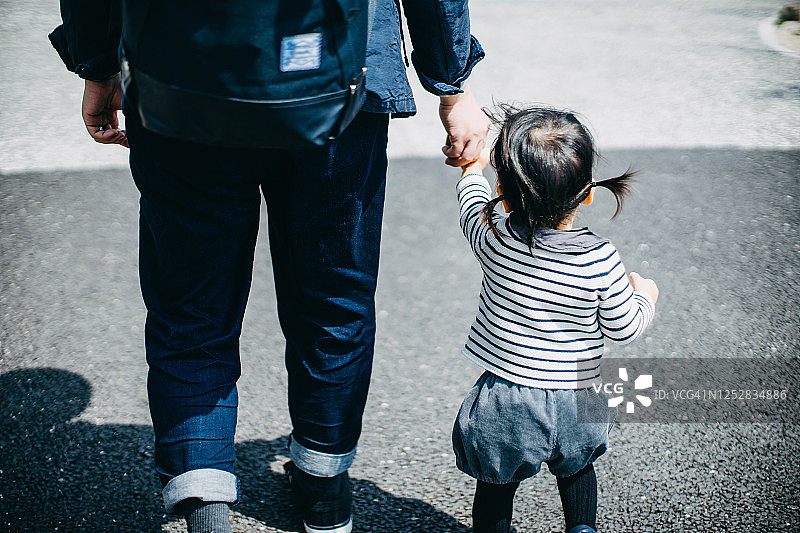 在一个阳光明媚的日子里，年轻的亚洲父亲牵着可爱的小女儿在户外的人行道上散步图片素材