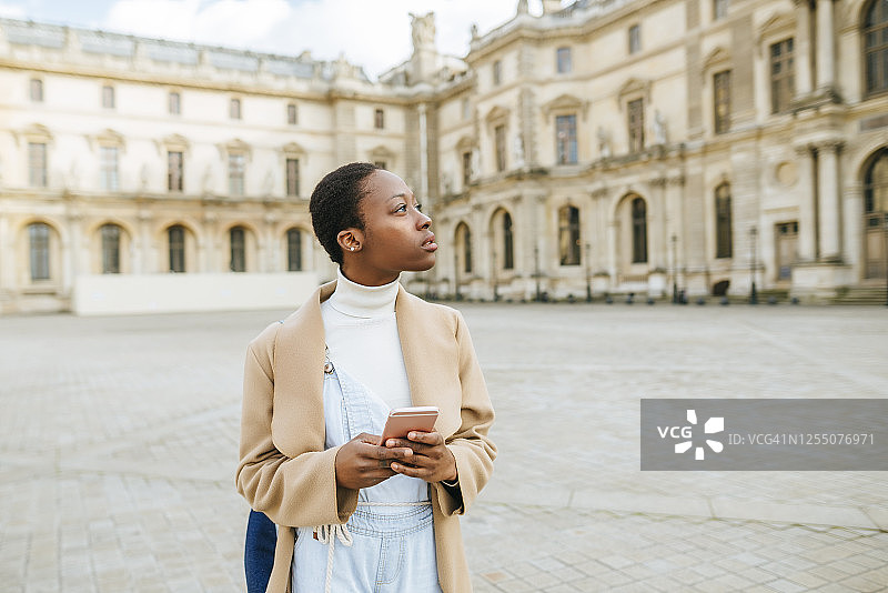 在法国巴黎，手持智能手机的体贴女人站在城市广场上，背景是一栋建筑图片素材