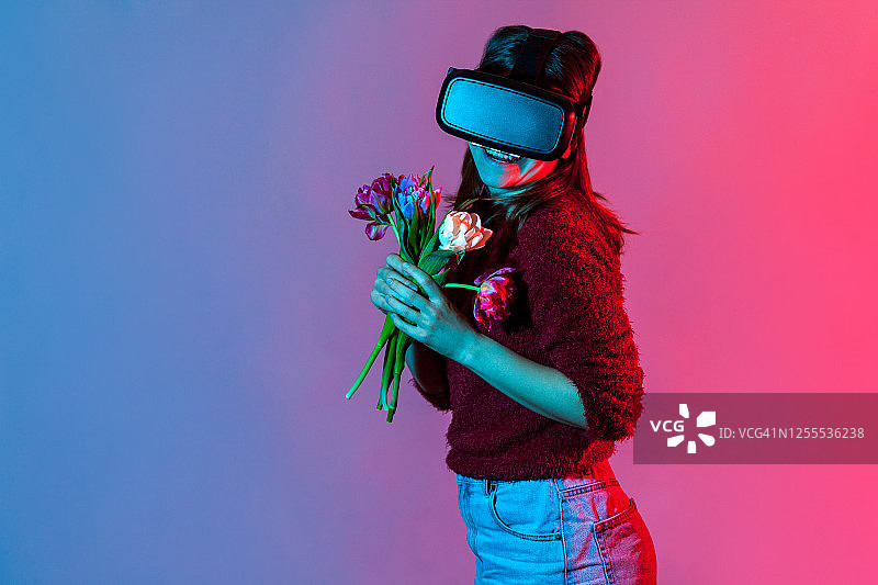 快乐可爱的女孩戴着VR眼镜戴着耳机捧着鲜花捧花，微笑着轻浮，玩着虚拟约会图片素材