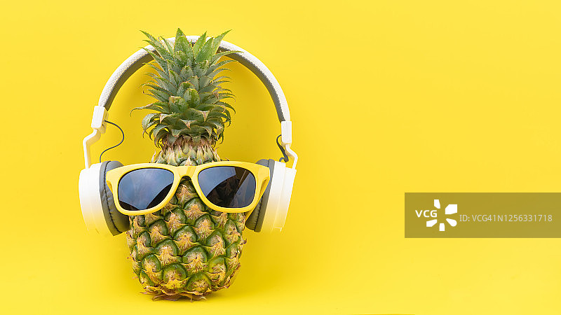 时尚菠萝戴着太阳镜和耳机在黄色背景下听音乐。热带夏季度假概念。夏日阳光明媚的热带岛屿海滩上图片素材