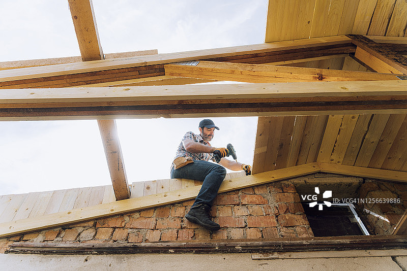 低角度的男性工人坐在木制屋顶上，向下看图片素材