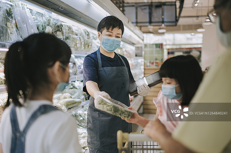 一位亚洲华人中年女性零售助理，建议并帮助一个家庭从超市的冷藏区选择蔬菜图片素材