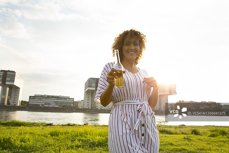 快乐的卷发女人拿着啤酒瓶，站在城市的河边图片素材