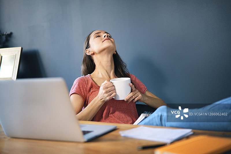 女性企业家拿着咖啡杯，在家庭办公室的办公桌上放松图片素材