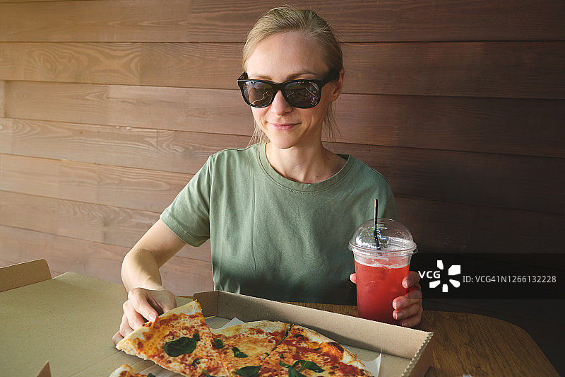 戴着墨镜的漂亮时尚女孩拿着一块素食披萨。意大利菜。木桌上的纸包装披萨。附近有软饮料或鸡尾酒。美味的午餐，小吃，快餐。图片素材