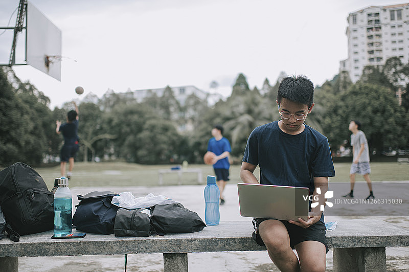 一个亚裔中国男孩在公园长椅上用笔记本电脑做作业，而他的朋友们在打篮球图片素材