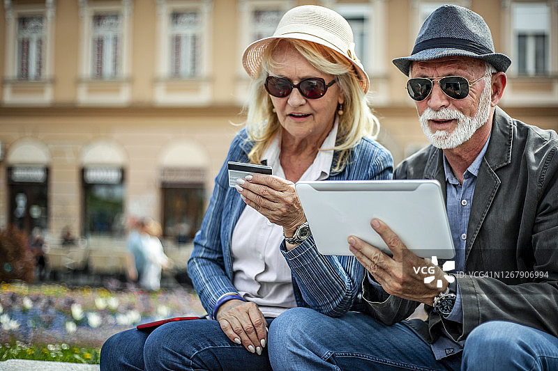 两个老人用平板电脑在网上购物——一对快乐的老夫妇坐在城市长廊上网购图片素材