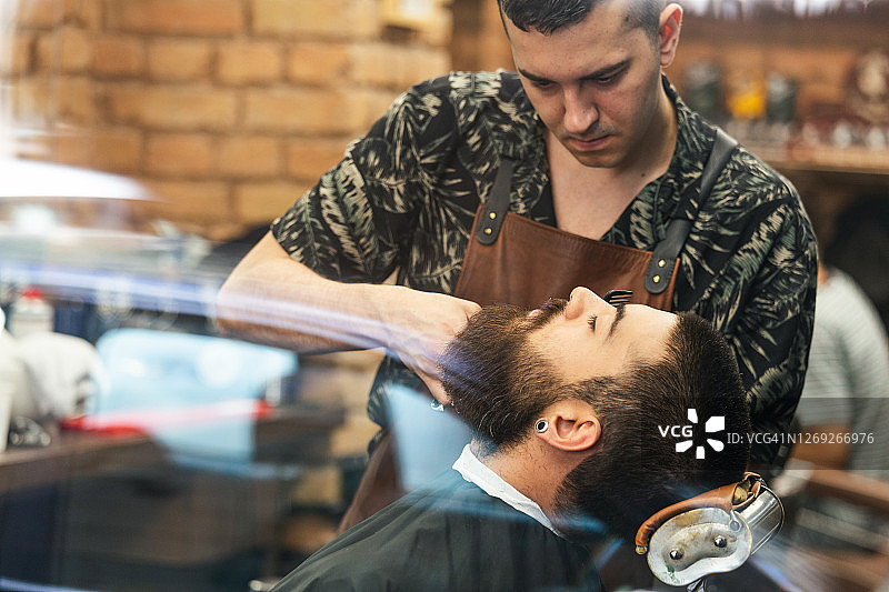 一名大胡子男子坐在理发店的扶手椅上，理发师用剪刀剪他的胡子。特写的理发师剪胡子的男人在理发店图片素材