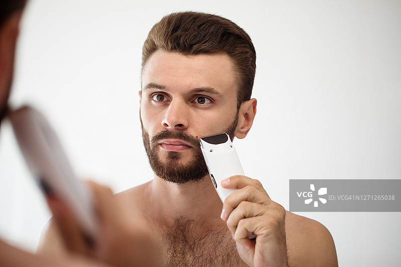 一个英俊的年轻人在浴室里刮胡子。一个时尚的裸胡子男人的肖像，正在检查他的脸在家里的镜子。图片素材