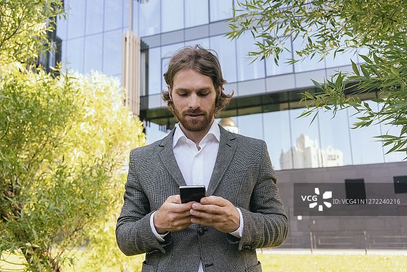 胡子英俊的商人使用智能手机，而站在办公大楼图片素材