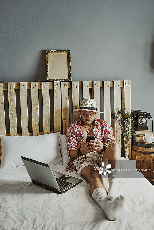 一个男人在酒店的床上用手机和笔记本电脑工作。图片素材