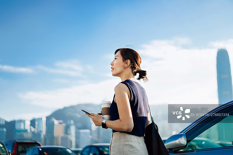 自信和专业的年轻亚洲商人走出她的车，拿着智能手机和一杯咖啡。在城市的映衬下仰望天空。商业上的概念图片素材