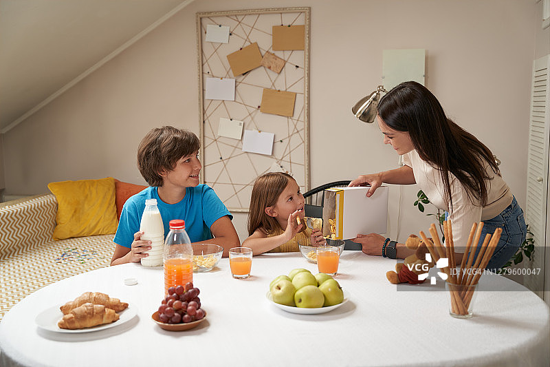 年轻的白人家庭，母亲和她的两个可爱的孩子微笑和交流，而坐在厨房桌子和在家吃早餐图片素材