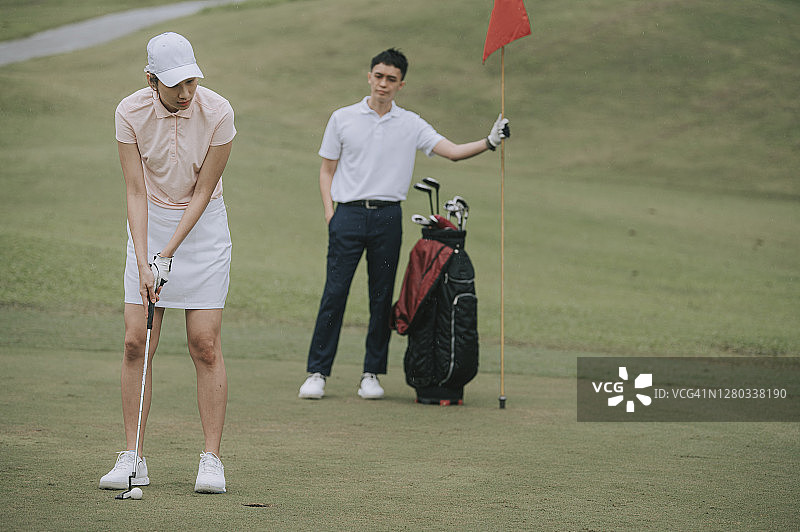 亚洲华人女子高尔夫球手在球场上与男伴举旗击球图片素材