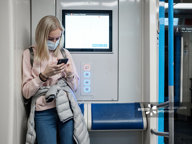地铁里，一名年轻女子拿着智能手机。乘客脸上戴着防护口罩。图片素材