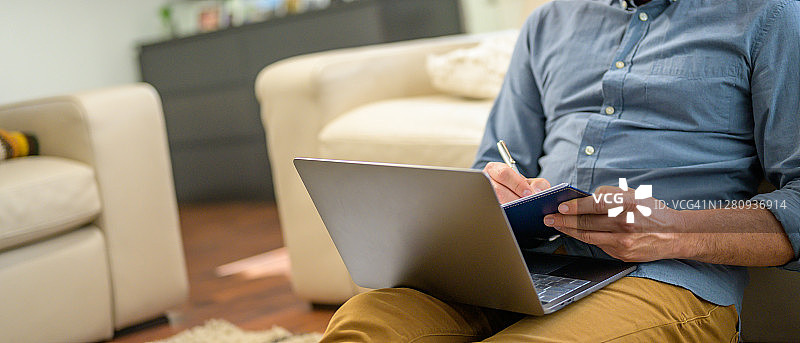 一个膝盖上放着笔记本电脑的男人正在写笔记。图片素材