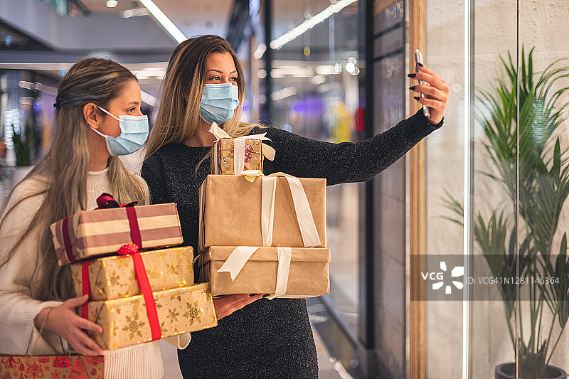 新冠肺炎疫情期间，两位美丽的女孩在商场享受圣诞购物时自拍。他们戴着防护口罩，以防止冠状病毒感染。图片素材