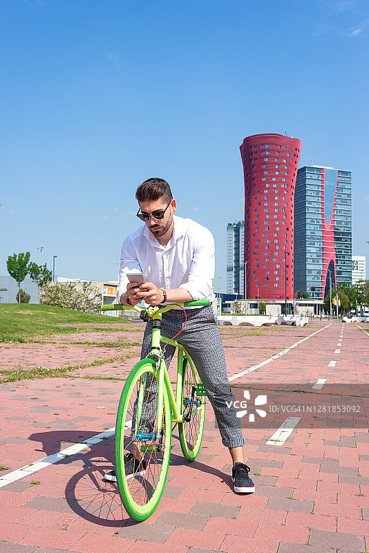 户外肖像英俊的年轻人用手机和固定齿轮自行车在街上。图片素材