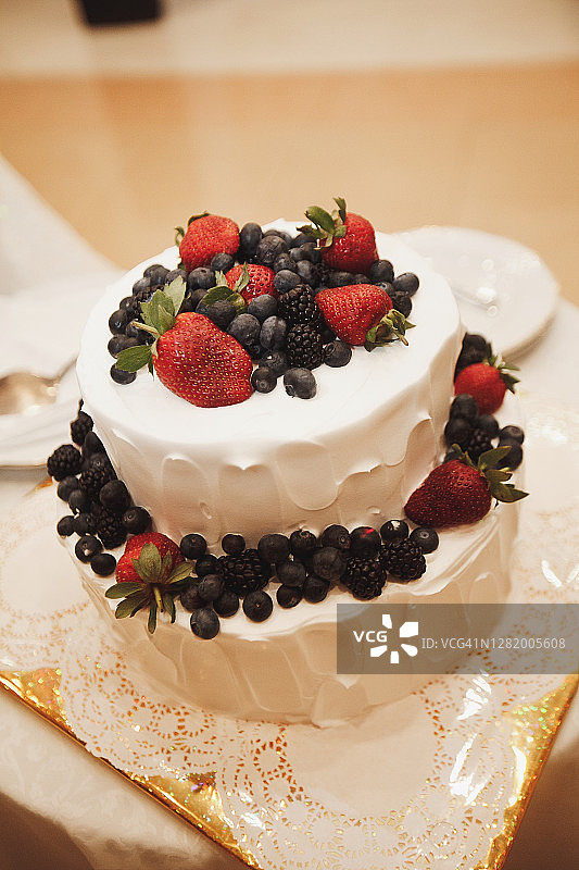一个优雅的两层婚礼蛋糕用浆果装饰在白色的木桌上。旁边是盘子，用来切片的餐具。图片素材