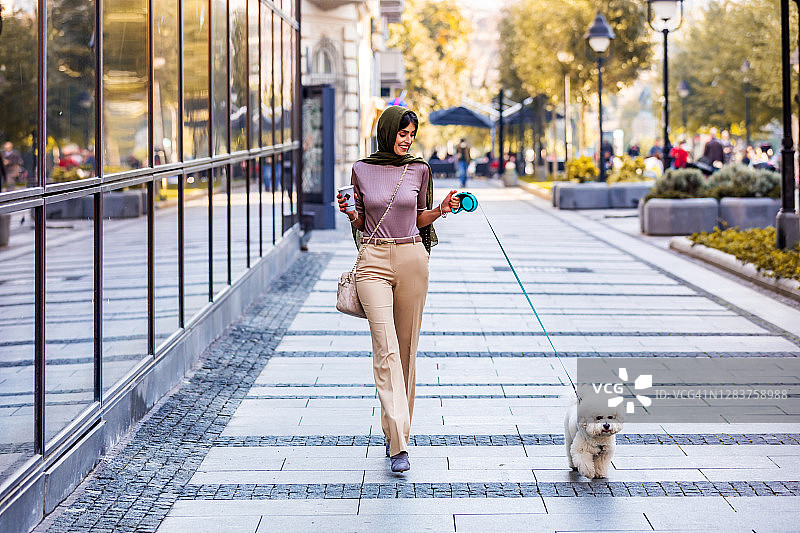 穆斯林成熟的妇女在温暖的衣服走她的小狗在办公楼附近的背景。图片素材