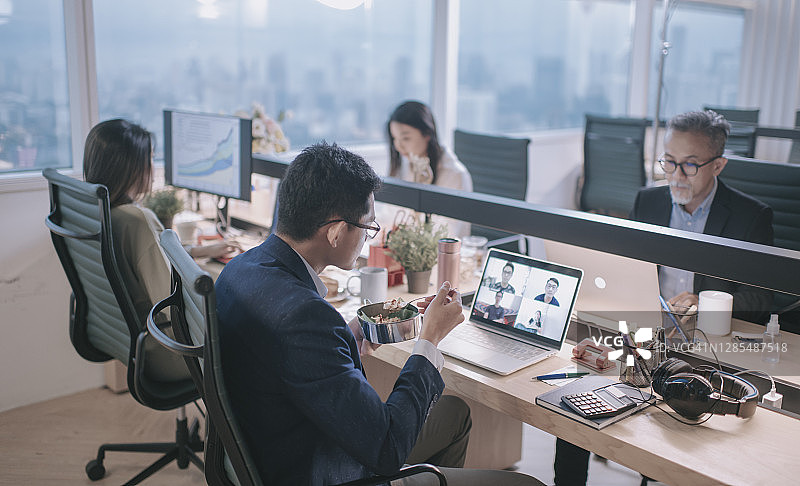 亚洲华人年轻商人视频电话在办公室工作，并在他的工作地点吃他的饭nasi lemak与他的客户在线商务会议图片素材