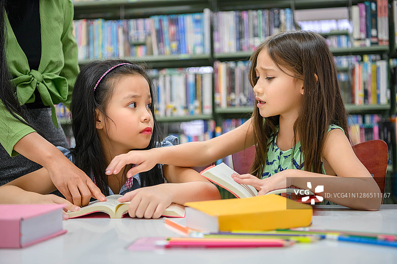 老师讲课时，两个可爱的女孩在图书馆看书时发生了冲突。人的生活方式和教育。在学校的概念中，年轻的友谊和孩子的关系。托儿所的主题图片素材