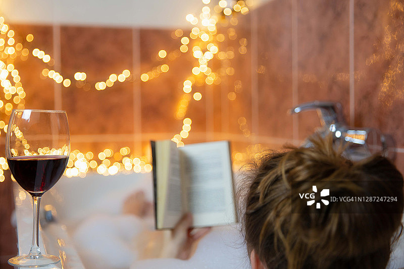 年轻美丽的女人在泡泡泡沫的浴缸里看书和喝红酒，用五颜六色的灯装饰，放松和水疗的概念图片素材