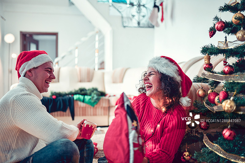 可爱的夫妇戴着圣诞帽，在树下喝着圣诞茶，笑着图片素材