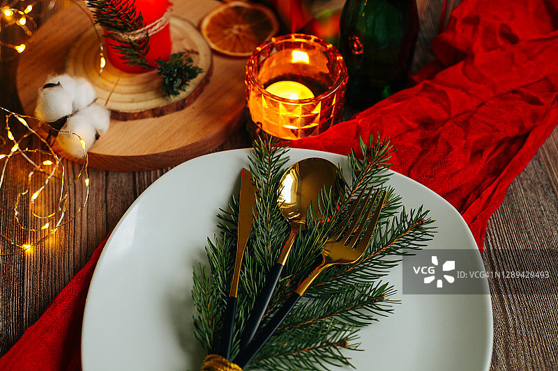 金色和黑色的刀叉，白色的盘子，红色的织物和木制的背景。松树枝、蜡烛和装饰品。为圣诞节和新年假期准备的节日餐桌。图片素材