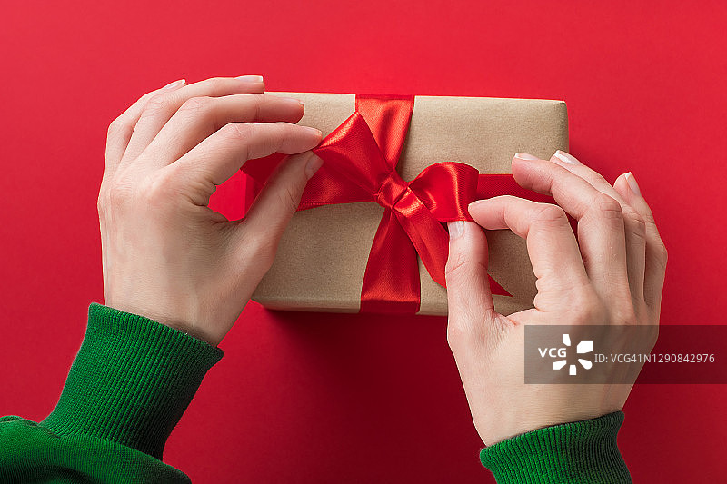 一个穿着绿色毛衣的女孩拿着一个漂亮的盒子，盒子的红色背景上用一条明亮的缎带系着一个惊喜。一个女人在她的节日打开礼物。快乐圣诞节的概念。图片素材
